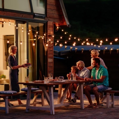 Mesa de madeira com copos e garrafas ao ar livre à noite com luzes decorativas e pessoas sentadas