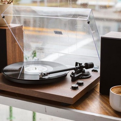 Gira-discos de madeira com vinil, colunas, café e vaso junto à janela