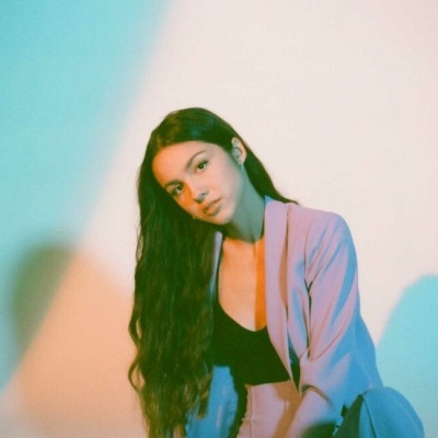 Mulher com casaco azul claro e top preto, cabelo longo e ondulado, sentada num fundo claro com iluminação colorida