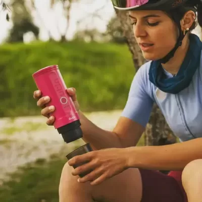 Mulher com capacete a servir bebida de uma garrafa térmica rosa para uma chávena preta ao ar livre