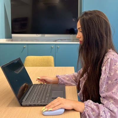 Portátil preto com teclado e rato branco numa secretária junto a uma jovem de casaco lilás floral em interior com armários azuis e televisão