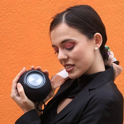 Mulher segura coluna de som portátil preta com painel metálico COOWE junto a parede laranja