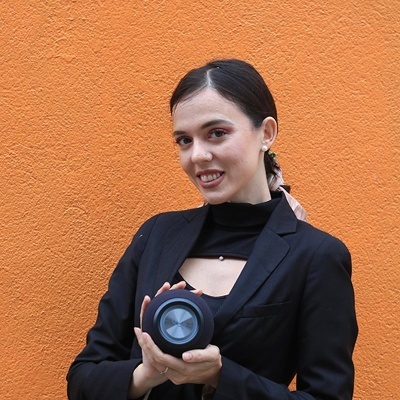Mulher com blazer preto segurando alto-falante portátil preto na frente de uma parede laranja