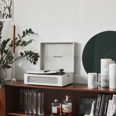 Aparelho de som branco Crosley num móvel de madeira com plantas, velas, garrafas e livros, com espelhos verdes ao fundo
