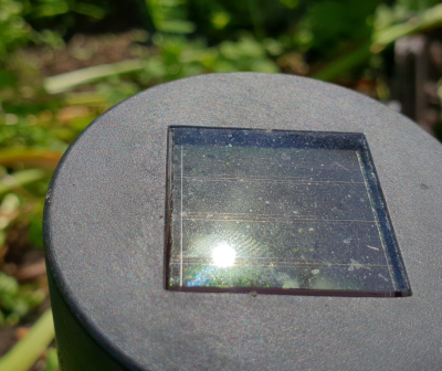 Pequeno painel solar quadrado sobre objeto cilíndrico cinzento escuro num jardim