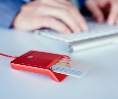 Leitor de cartões vermelho com cartão branco inserido ao lado de teclado branco e mãos desfocadas ao fundo