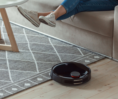 Aspirador robot preto em chão de madeira junto a tapete cinza com padrão branco e sofá bege.