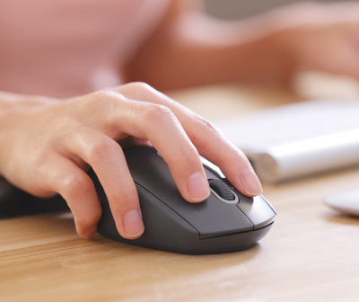 Mão usando rato de computador preto na mesa de madeira clara com portátil ao fundo.