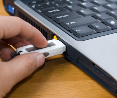 Mão a inserir pen USB branca num portátil preto e prateado sobre mesa de madeira