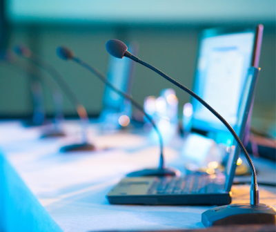 Microfones com bases cinzentas e espuma preta em mesa branca com computadores e copos de água