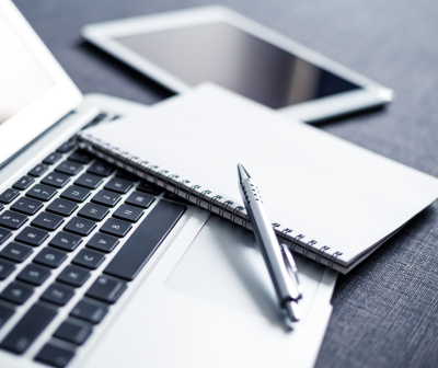 Notebook prata com teclado preto, caderno de espiral branco, caneta metálica prateada e tablet preto sobre tecido cinzento