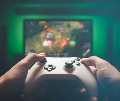 Comando de videojogo branco com botões coloridos nas mãos em frente a ecrã de videojogo