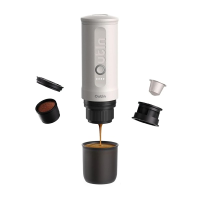 Máquina de café portátil a preparar café para um copo preto, com cápsulas e pó de café à volta