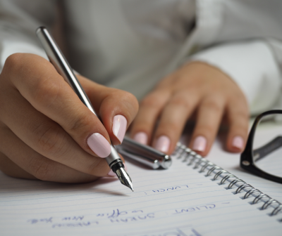 Mão a escrever com caneta metálica num caderno de espiral ao lado de óculos pretos