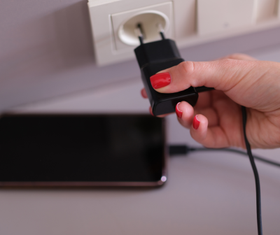 Mão segurando carregador preto a ser ligado a tomada branca com smartphone preto na superfície