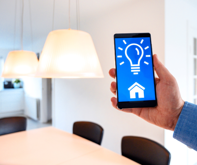 Pessoa segura telemóvel com app de luz, mostrando ícones de lâmpada e casa num ambiente de sala de jantar moderna