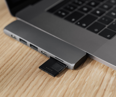 Adaptador USB-C cinzento com cartão SD ligado a portátil prateado em mesa de madeira
