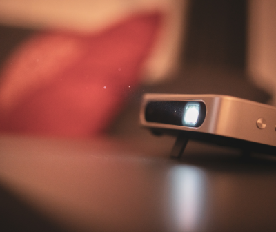 Projetor compacto cinza com luz branca sobre superfície escura