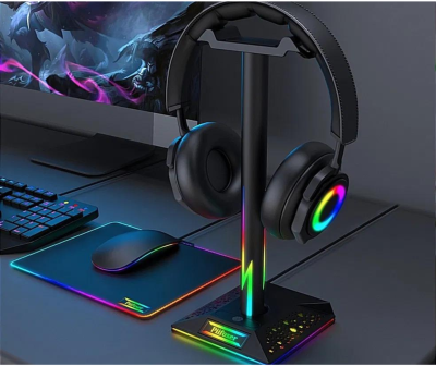 Suporte para auscultadores preto com iluminação RGB ao lado de auscultadores e acessórios de gaming numa mesa.