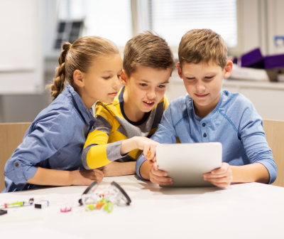 Três crianças a usar tablet branco numa mesa com objetos eletrónicos