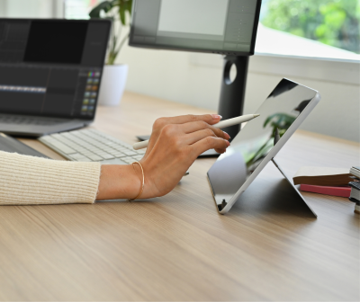 Mão usando caneta digital num tablet prateado numa mesa de escritório