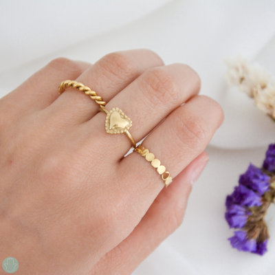 Anel duplo dourado com coração sólido e padrão torcido em mão sobre fundo claro com flores secas