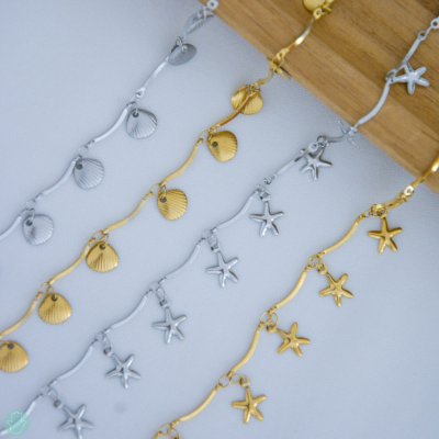 Pulseiras de metal dourado e prateado com pendentes de estrela do mar e concha penduradas em suporte de madeira