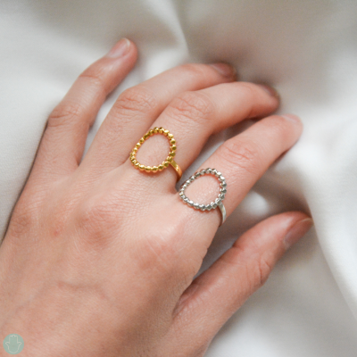 Anéis dourado e prateado com laço aberto, de esferas, usados na mão esquerda sobre tecido branco.