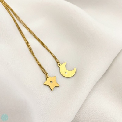 Duas correntes douradas com pendentes em forma de estrela e lua com brilho no centro sobre tecido branco