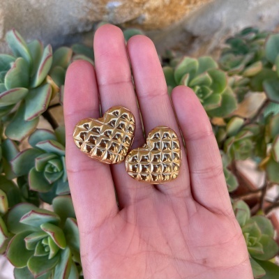 Dois corações dourados com relevo quadriculado numa mão com fundo de plantas suculentas.