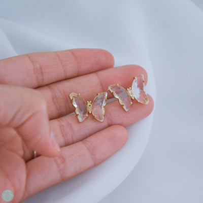 Brincos de borboleta dourados com detalhe transparente numa mão sobre tecido branco