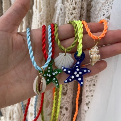 Cinco pulseiras coloridas com pendentes de conchas e estrelas do mar em mão sobre fundo de tecido branco trançado