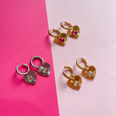 Três pares de brincos de argola com corações pendentes, dourados e prateados, em fundo rosa e branco