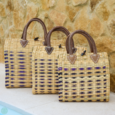 Três cestas de palha com alças em couro castanho sobre superfície branca e parede de pedra amarela