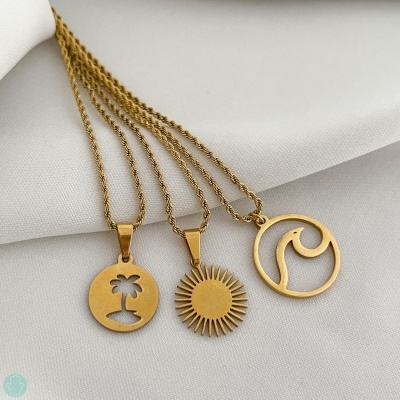 Três colares dourados com pendentes de palmeira, sol e onda sobre tecido branco.