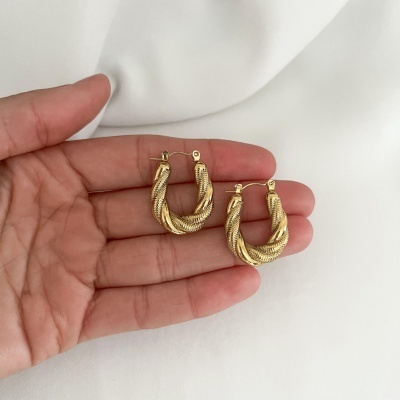 Par de brincos dourados torcidos em forma de argola na mão sobre tecido branco