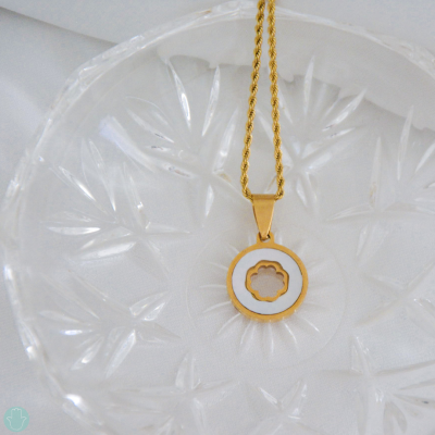 Colar dourado com pendente redondo branco e floral, sobre prato de vidro texturizado