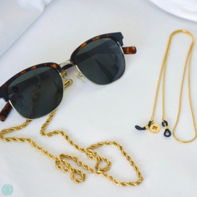Óculos de sol com armação preta e castanha e correntes douradas sobre superfície branca