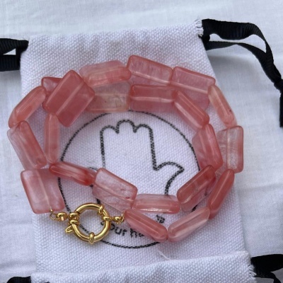 Pulseira rosa com fecho dourado sobre saco de pano branco com símbolo de mão