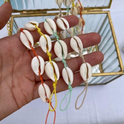 Pulseiras com conchas brancas em cordões coloridos seguradas em mão junto a caixa de vidro dourada