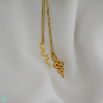 Dois colares dourados com pendentes em forma de serpente sobre tecido branco.