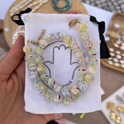 Colar de contas de vidro coloridas sobre saco branco com desenho de mão e texto É EMMA