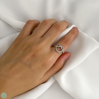 Anel prateado com pedras em duas formas circulares entrelaçadas na mão sobre tecido branco