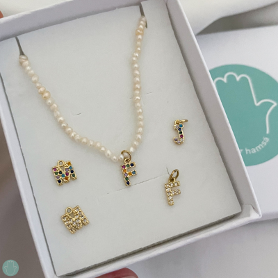 Conjunto de colar de pérolas com pendente e vários pendentes de letras dourados com pedras coloridas e brancas numa caixa branca