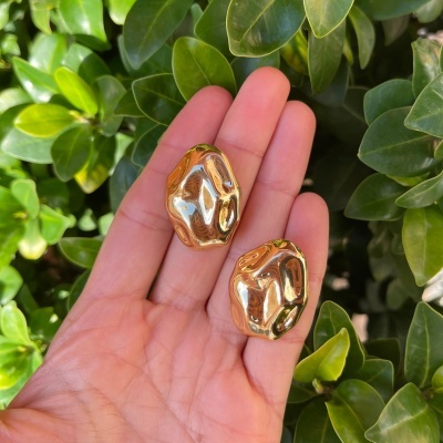 Brincos dourados com design orgânico, mão segurando, fundo de folhas verdes