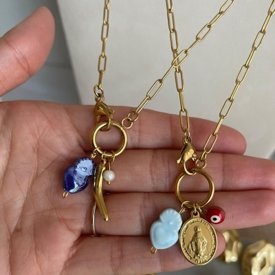 Dois colares dourados com vários pendentes, incluindo conta azul, pérola, medalha dourada e conta vermelha, no fundo neutro.