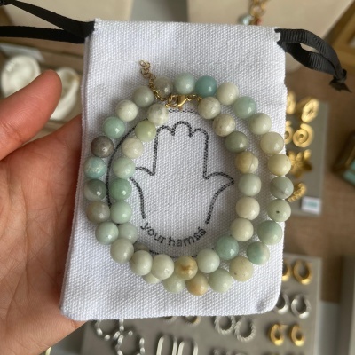 Pulseira de contas coloridas com fecho dourado sobre bolsa de tecido branco com mão de hamsa