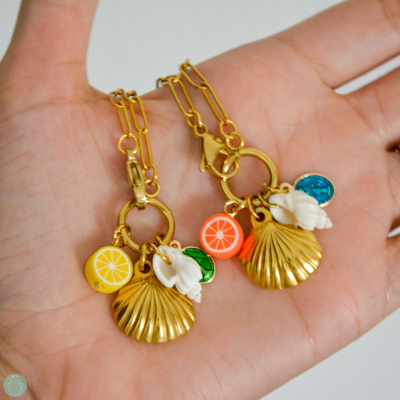 Dois colares dourados com pendentes de conchas, frutas e folha colorida na mão.
