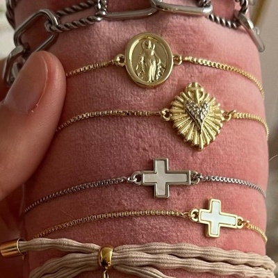 Cinco pulseiras delicadas douradas e prateadas com pendentes de medalha, folha e cruz, sobre suporte cor de rosa.
