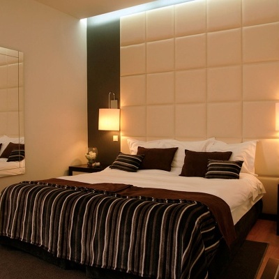 Quarto com cama de casal com cabeceira estofada creme e roupa de cama em castanho e riscas.